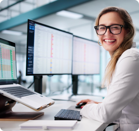 Smiling professional wearing glasses working at a desk with multiple computer monitors displaying data in a modern office environment.
